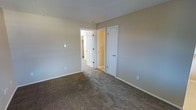 This single-floor residential space offers a clean and functional layout designed for comfortable everyday living. The interior is finished with neutral beige-toned walls that create a calm, understated ambiance throughout. The flooring features wall-to-wall carpet in a warm brown tone, adding softness and a sense of coziness underfoot.

The room visible in the space is generously proportioned, providing ample room for furnishings and movement. A textured popcorn ceiling adds a classic residential character overhead. Two white paneled doors are prominently featured — one leading to an adjacent hallway or common area, illuminated by warm yellow-toned lighting, and a second door that likely accesses a closet, offering convenient built-in storage. White baseboards frame the perimeter of the room, providing a crisp contrast against the neutral walls and carpet.

The overall layout suggests a well-connected interior, with the hallway beyond offering access to additional areas of the home. The space is bright, open, and move-in ready, presenting a straightforward and practical residential environment suited for a variety of uses and furnishing styles.