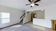 This two-story home offers a thoughtfully designed layout that balances open living spaces with practical functionality. The main level centers around a spacious living area finished with plush carpeting and light tile flooring, creating a comfortable and welcoming atmosphere. A ceiling fan with a built-in light fixture is mounted overhead, promoting air circulation and adding a classic touch to the room.

A staircase with dark wood railings rises gracefully from the main living area, serving as a central architectural feature that connects the two floors. Adjacent to the living space, a kitchen counter or breakfast bar is partially visible, suggesting an open-concept flow between the living and kitchen areas. The kitchen area appears to transition smoothly from the tiled entryway, offering a practical and accessible cooking space.

Natural light filters in through a window near the front entry, brightening the main level and enhancing the home's airy feel. The neutral color palette throughout — from the light walls to the soft flooring — provides a versatile backdrop suited to a variety of interior styles. The upper floor, accessible via the interior staircase, extends the home's living capacity, offering additional private spaces above the main level.
