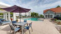 Located at 10010 Skinner Lake Dr in Jacksonville, Florida, this expansive outdoor amenity space centers around a generously sized swimming pool with crystal-clear turquoise water. The pool deck is finished with tasteful paved tile and is lined with multiple lounge chairs and seating arrangements, offering ample space for relaxation and recreation. A classic white pergola structure anchors one end of the deck, providing a shaded gathering area accented with additional seating. Safety equipment, including a life ring, is conveniently mounted along the adjacent building exterior, which features a distinctive red tile roofline and soft blue-grey stucco finish. Lush mature trees frame the perimeter, enhancing the sense of natural seclusion while manicured hedgerows define the boundaries of the space. The layout extends toward a waterfront area along Skinner Lake, where a dock structure and additional outdoor seating areas offer direct lakeside access and sweeping water views. Umbrella-shaded tables and chairs provide a comfortable setting for outdoor dining or leisure near the lake's edge. Multi-story residential buildings are visible in the background, complementing the well-maintained grounds. The overall ambiance is bright, open, and inviting, ideal for enjoying Florida's warm climate year-round.
