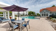Located at 10010 Skinner Lake Dr in Jacksonville, Florida, this expansive outdoor amenity space centers around a generously sized swimming pool with crystal-clear turquoise water. The pool deck is finished with tasteful paved tile and is lined with multiple lounge chairs and seating arrangements, offering ample space for relaxation and recreation. A classic white pergola structure anchors one end of the deck, providing a shaded gathering area accented with additional seating. Safety equipment, including a life ring, is conveniently mounted along the adjacent building exterior, which features a distinctive red tile roofline and soft blue-grey stucco finish. Lush mature trees frame the perimeter, enhancing the sense of natural seclusion while manicured hedgerows define the boundaries of the space. The layout extends toward a waterfront area along Skinner Lake, where a dock structure and additional outdoor seating areas offer direct lakeside access and sweeping water views. Umbrella-shaded tables and chairs provide a comfortable setting for outdoor dining or leisure near the lake's edge. Multi-story residential buildings are visible in the background, complementing the well-maintained grounds. The overall ambiance is bright, open, and inviting, ideal for enjoying Florida's warm climate year-round.
