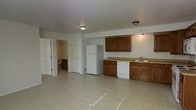 This single-floor space offers an open layout that combines the kitchen and living area into one cohesive environment. The kitchen is equipped with warm-toned wood cabinetry providing ample storage, a light-colored countertop, a stainless steel sink, a white refrigerator, and a dishwasher. The cabinetry is arranged along one wall, creating an efficient and functional workspace. Overhead, flush-mount light fixtures illuminate the space with a neutral, even glow, complementing the white walls and light tile flooring that extends throughout the main area. The tile flooring adds a clean, easy-to-maintain surface that ties the open space together. Two doorways are visible, leading to additional rooms — one of which appears to open into a bathroom or secondary space with its own cabinetry and fixtures, suggesting practical utility. The overall layout is straightforward and functional, with a neutral color palette of whites and warm browns that creates a simple, livable atmosphere. The open design between the kitchen and the adjoining area allows for flexible use of the space.