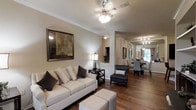 This single-story space showcases an open-concept design that seamlessly connects the living and dining areas, creating a cohesive and inviting atmosphere. The living area is anchored by a plush, cream-toned sofa accented with decorative pillows in neutral and charcoal hues, complemented by a stylish side table and an elegant table lamp. A large framed artwork adds a sophisticated focal point to the warm beige walls, which are finished with crisp crown molding. A ceiling fan with an integrated light fixture provides both comfort and ambient illumination overhead.

Rich hardwood flooring flows continuously throughout the space, adding warmth and visual continuity. A sleek accent chair in dark upholstery bridges the living and dining zones, while patterned ottomans offer additional flexible seating. The dining area at the far end features a round table surrounded by upholstered chairs, illuminated by a ceiling-mounted track light fixture. A window in the dining space allows natural light to filter in, brightening the entire open layout. The overall ambiance is modern yet comfortable, with a carefully curated neutral palette and layered lighting.