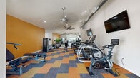 Located at 5150 W Eugie Ave in Glendale, Arizona, this fitness center offers a well-equipped, single-floor workout facility designed to support a full range of exercise routines. The space features vaulted ceilings that lend an open, airy feel, complemented by recessed lighting and multiple large ceiling fans that promote air circulation throughout. An exposed HVAC duct runs prominently along one wall, adding an industrial design element to the otherwise clean, modern aesthetic.

The cardio zone is well-stocked with treadmills, elliptical trainers, stair climbers, and stationary bikes, all arranged in an organized row for easy access. A wall-mounted flat-screen television is positioned for viewing during workouts. The strength training area includes a multi-function cable machine, a weight bench, and a dedicated free weights section featuring a full dumbbell rack positioned beneath large windows. The windows allow natural light to filter in and provide views of the surrounding outdoor area.

An accent wall painted in a warm amber-orange tone adds visual interest, while motivational fitness artwork adorns the walls. The flooring consists of a bold chevron-patterned carpet in shades of orange, blue, and gray, tying the energetic design together. This fitness center provides a comprehensive and inviting environment for residents to pursue their health and wellness goals.