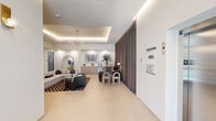 This single-floor space presents a thoughtfully designed lobby and communal lounge area that combines modern aesthetics with functional layout. The entrance corridor welcomes visitors with large-format light tile flooring, recessed and cove lighting set within a tray ceiling, brass wall sconces, and floor-to-ceiling glass entry doors that draw in natural light and views of the surrounding exterior. An elevator and stairwell access are conveniently situated near the entry, alongside potted greenery that adds a welcoming touch.

The lounge area is anchored by a generous sectional sofa in light grey, complemented by a bold black-and-pink checkered area rug and a glass-top coffee table. Accent seating includes dark upholstered chairs and sculptural side tables, creating distinct conversation zones. A striking multi-globe brass chandelier suspended from a recessed tray ceiling serves as a focal point overhead, enhanced by continuous cove lighting that lends the space a warm, ambient glow.

Built-in wood-toned credenzas line the walls, topped with curated décor including potted plants, ceramic vessels, and framed artwork. A wall-mounted flat-screen display is integrated into a niche above one credenza. Floor-to-ceiling draped curtains divide the lounge from an adjacent meeting or workspace area, offering flexible privacy. The overall ambiance is sophisticated, clean, and contemporary throughout.