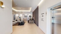 This single-floor space presents a thoughtfully designed lobby and communal lounge area that combines modern aesthetics with functional layout. The entrance corridor welcomes visitors with large-format light tile flooring, recessed and cove lighting set within a tray ceiling, brass wall sconces, and floor-to-ceiling glass entry doors that draw in natural light and views of the surrounding exterior. An elevator and stairwell access are conveniently situated near the entry, alongside potted greenery that adds a welcoming touch.

The lounge area is anchored by a generous sectional sofa in light grey, complemented by a bold black-and-pink checkered area rug and a glass-top coffee table. Accent seating includes dark upholstered chairs and sculptural side tables, creating distinct conversation zones. A striking multi-globe brass chandelier suspended from a recessed tray ceiling serves as a focal point overhead, enhanced by continuous cove lighting that lends the space a warm, ambient glow.

Built-in wood-toned credenzas line the walls, topped with curated décor including potted plants, ceramic vessels, and framed artwork. A wall-mounted flat-screen display is integrated into a niche above one credenza. Floor-to-ceiling draped curtains divide the lounge from an adjacent meeting or workspace area, offering flexible privacy. The overall ambiance is sophisticated, clean, and contemporary throughout.
