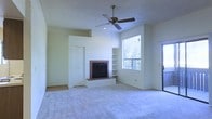 This single-story space centers around a well-appointed living area that combines functional design with comfortable finishes. The room features a wood-trimmed fireplace set against a clean white wall, serving as a natural focal point. Flanking the fireplace are built-in shelving units, offering practical display and storage options. Soft, neutral-toned carpeting covers the floor, contributing to a warm and inviting atmosphere throughout the space.

Vaulted or elevated ceilings add a sense of openness and volume to the room, enhanced by a ceiling fan with a light fixture that provides both airflow and ambient illumination. A large window positioned beside the fireplace wall floods the interior with natural light and frames views of surrounding greenery, creating a pleasant connection to the outdoors.

A door adjacent to the window suggests access to an outdoor area, further extending the living space. A closet with white panel doors is visible along one wall, providing convenient built-in storage. The overall color palette — composed of whites and soft neutrals — lends the space a clean, airy feel well-suited to a variety of interior styles.