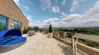 This well-appointed space offers a thoughtfully curated mix of amenities across its single floor. The standout feature is a spacious rooftop terrace finished with large-format stone tiles, furnished with multiple seating arrangements and accented by potted evergreen topiaries. The terrace is enclosed by metal railings, offering sweeping panoramic views of the surrounding cityscape, treetops, and distant rolling hills beneath an open sky.

Inside, a fitness area occupies a bright, windowed corner room with dark hardwood-style flooring and roller shades. Floor-to-ceiling windows wrap around the space on multiple sides, flooding it with natural light and framing the same impressive outdoor views. Equipment includes a rowing machine, a stationary spin cycle, a balance ball, and a stretching board, complemented by a granite-topped worktable and a cushioned accent chair.

Adjacent to the fitness area is a lounge or common area featuring a striking textured wave-pattern accent wall with a wall-mounted flat-screen television. The room is furnished with upholstered seating and connects to a corridor with recessed lighting and wood-toned flooring, creating a cohesive and modern interior aesthetic throughout.