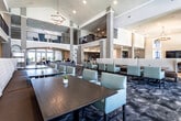This elegantly designed community clubhouse or leasing center offers a refined and welcoming atmosphere across a single, well-appointed floor. The centerpiece is a dramatic two-story grand lobby with soaring vaulted ceilings, decorative chandeliers, and arched architectural details that create an open, airy feel. The main lounge area features a bold circular sectional sofa in a navy geometric pattern, surrounded by accent seating on a richly patterned carpet, anchoring the space with a distinct sense of style.

Adjacent to the lobby, a dining area boasts double-height ceilings lined with expansive windows that flood the room with natural light and offer outdoor views. Dark wood dining tables paired with upholstered chairs provide a comfortable, social setting, complemented by a statement ring chandelier overhead.

A full catering-style kitchen features dark espresso cabinetry, quartz countertops, a blue tile mosaic backsplash, and stainless steel appliances including double wall ovens, a refrigerator, and a dishwasher — ideal for events and gatherings.

A dedicated reception area with a herringbone-accent wall serves as the leasing office, while wide parquet-patterned flooring, curated wall art, and lush greenery accents tie the entire space together with a polished, hospitality-inspired aesthetic.