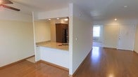 This single-floor space is designed with an open and functional layout that promotes a seamless flow between its main living areas. The kitchen is partially enclosed by a half-wall partition, creating a pass-through breakfast bar topped with light granite countertops that add a touch of elegance. Inside the kitchen, dark espresso-toned cabinetry provides strong visual contrast against the lighter walls and counters, while recessed lighting illuminates the workspace. A black refrigerator anchors the kitchen, contributing to the cohesive, contemporary aesthetic.

Warm hardwood flooring extends throughout the space, tying the kitchen and living areas together with a consistent, inviting tone. The open corridor leading toward the living area benefits from natural light streaming in through a window at the far end, brightening the interior and creating an airy feel. Clean white walls and neutral finishes throughout keep the space feeling fresh and versatile. Baseboard heating is visible near the window, and the overall layout is straightforward and practical, making it well-suited for everyday living.