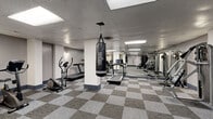 This fitness center offers a spacious, single-floor layout designed to accommodate a comprehensive range of exercise equipment and workout styles. The floor is finished with a distinctive geometric-patterned carpet tile in neutral grey and blue tones, lending the space a clean, functional aesthetic throughout.

The main workout area is organized to maximize usability, with cardio equipment—including treadmills, elliptical trainers, and stationary bikes—arranged along one side. Structural columns punctuate the open floor plan, one of which serves as the anchor point for a suspended Venum heavy punching bag, adding a dedicated boxing and functional training element to the space.

A strength training section features a rack of dumbbells in varying weights, a weight bench, and a cable machine system, offering users versatile resistance training options. Additional multi-use strength machines line the perimeter, while a wall-mounted flat-screen television provides entertainment during workouts.

The ceiling features a standard drop tile grid with integrated recessed fluorescent panel lighting, ensuring even illumination across the entire space. Grey-painted walls and support columns contribute to the neutral, no-frills atmosphere. A staircase at the rear of the space provides access to adjoining areas of the building.
