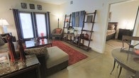 This single-floor space features a thoughtfully arranged living area that balances functionality with distinctive style. The flooring throughout is polished concrete, providing a sleek, low-maintenance foundation that complements the warm tones of the furnishings. Natural light enters through a large window dressed with horizontal blinds and dark curtain panels, illuminating the room and creating a bright, welcoming atmosphere.

The living area is anchored by a neutral-toned sectional chaise paired with a zebra-print accent pillow, adding a bold decorative touch. A low-profile coffee table and a vibrant red shag area rug define the central seating zone, injecting warmth and texture into the space. An armchair with an orange accent pillow occupies a corner near the window, offering an additional seating option.

Two tall, dark-wood leaning ladder shelves serve as a standout architectural and storage feature along one wall, displaying decorative objects, artwork panels, and greenery. A narrow console table sits between the shelves, enhancing the gallery-like arrangement. The overall ambiance is casual yet curated, with an eclectic mix of textures and earthy tones throughout.