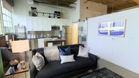 This single-floor loft-style space blends industrial aesthetics with modern comfort in an open-concept layout. Soaring ceilings with exposed ductwork and structural elements give the space a distinctly urban character, while the open floor plan allows the living and kitchen areas to flow seamlessly into one another.

The kitchen is equipped with stainless steel appliances, including a refrigerator and microwave, and features white shaker-style cabinetry paired with a subway tile backsplash. A compact kitchen island provides additional prep and dining space, making the kitchen both functional and visually appealing.

The living area is anchored by a charcoal gray sofa accented with layered throw pillows and a cozy blanket, creating an inviting and casual atmosphere. A sliding barn door adds a stylish architectural detail while offering a practical room divider, contributing to the loft's contemporary-industrial design language.

Polished concrete-style flooring runs throughout, tying the space together cohesively. The combination of exposed mechanical elements overhead, clean white cabinetry, and modern furnishings creates a balanced aesthetic that feels both functional and thoughtfully designed.