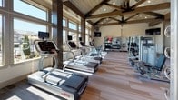 This fitness center is a standout facility defined by its striking architectural character and comprehensive equipment offerings. The space is anchored by soaring vaulted ceilings adorned with exposed wood beam trusses and a wood-paneled ceiling, lending a warm yet refined aesthetic to the workout environment. Large, floor-to-ceiling windows line the walls, flooding the interior with abundant natural light and offering open views of the surrounding outdoor areas, including a pool and landscaped grounds.

The flooring throughout consists of wide-plank wood-tone vinyl, providing a clean and durable surface suited to high-traffic fitness use. The cardio zone is generously equipped with a full row of treadmills, elliptical trainers, upright and recumbent stationary bikes, and a rowing machine — all positioned to take advantage of the natural light and outdoor views. Mounted flat-screen televisions are strategically placed for entertainment during workouts.

The strength training area includes a multi-station cable and weight machine, free weights, medicine balls, and additional resistance equipment neatly organized along the walls. A separate adjoining room, accessible through glass-paneled doors, provides additional workout space. The overall ambiance is bright, airy, and motivating — combining thoughtful design with a well-rounded selection of fitness amenities.