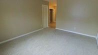 This single-floor space presents a clean and functional layout with a neutral color palette throughout. The room visible in the image features wall-to-wall carpeting in a light, speckled tone that adds warmth and softness underfoot. The walls are finished in a muted taupe hue, complemented by crisp white baseboards that provide a subtle contrast and a sense of clean definition along the floor line.

A louvered bifold closet door is positioned along one wall, offering practical storage access while contributing a classic architectural detail to the room. A hallway visible in the background connects to an additional area, where a second louvered closet door can be seen, suggesting consistent storage solutions throughout the space. The flooring in the hallway transitions to a harder surface, providing a natural visual distinction between spaces.

The layout appears open and straightforward, allowing for flexible furniture arrangement. The absence of clutter and the neutral finishes give the space a blank-canvas quality, making it highly adaptable to a variety of uses and décor styles. Overall, the space conveys a modest, move-in-ready character with consistent, practical finishes.