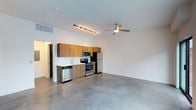 This single-floor space showcases a clean, modern aesthetic defined by an open-concept layout that seamlessly integrates the kitchen and living areas. Polished concrete floors extend throughout, lending an industrial character that pairs well with the crisp white walls and elevated ceilings. The kitchen is positioned along one wall and features warm wood-toned cabinetry, a full suite of stainless steel appliances including a refrigerator, range, microwave, and dishwasher, and a single-basin sink. Above, a track lighting fixture provides focused illumination over the kitchen area, while a ceiling fan with a sleek, low-profile design serves the open living space, contributing to both comfort and airflow. The generous open floor plan allows for flexible furniture arrangement in the living area, making the space highly adaptable. A hallway visible near the kitchen suggests access to additional rooms beyond the main living area. The overall ambiance is contemporary and minimalist, with the combination of industrial flooring, modern fixtures, and neutral tones creating a cohesive and versatile environment.