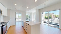 This modern single-floor unit offers five bedrooms, two bathrooms, and a thoughtfully designed open-concept layout. The kitchen and living room form the heart of the space, anchored by a sleek white island with a built-in sink and matte black faucet. Full-height cabinetry in a crisp white finish lines the kitchen walls, complemented by stainless steel appliances including a refrigerator, range, and microwave. Recessed lighting illuminates the space, which flows seamlessly into the living area via light wood flooring found throughout. Large floor-to-ceiling windows and a sliding glass door flood the living area with natural light while offering open views of the surrounding cityscape and neighborhood. The bedrooms are well-proportioned, each featuring the same light hardwood floors and clean white walls. Several bedrooms benefit from oversized windows with expansive urban views. One bedroom includes a stylish barn-style sliding door. A dedicated closet provides built-in wire shelving for organized storage. The hallway connects the rooms with a clean, minimalist aesthetic, featuring recessed ceiling vents and lighting. Both bathrooms are finished with wood-look flooring, white tile walk-in showers with matte black hardware, and clean, simple cabinetry. The overall ambiance of the space is bright, airy, and contemporary.