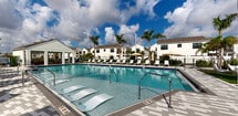 Located at 9145 Flying Eagle Dr in Fort Myers, Florida, this single-level outdoor amenity space offers a well-appointed community retreat designed to complement the Florida lifestyle. At the heart of the area is a generously sized resort-style swimming pool with crystal-clear blue water, surrounded by an expansive paver deck lined with white lounge chairs and shade umbrellas — ideal for sunbathing and relaxation.

A covered pavilion anchors the space architecturally, featuring a pitched white roof supported by clean columns. Beneath it sits a dining area with a white table and wicker chairs, along with an adjacent outdoor grill station, making it well-suited for outdoor entertaining. The pavilion opens directly to the pool deck, creating a seamless indoor-outdoor flow.

Beyond the pool, a manicured lawn area provides open green space for recreation, including a cornhole game setup framed by tall palm trees. A separate, more intimate seating nook features modern butterfly chairs arranged on a turf pad surrounded by tropical landscaping — perfect for quiet gathering.

Throughout the space, lush plantings including palms, ornamental grasses, shrubs, and mulched garden beds add tropical character and visual depth. The entire area is enclosed by a decorative iron fence, providing privacy while maintaining an open, airy ambiance under the sunny Southwest Florida sky.