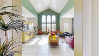 Located at 11400 Edgemere Blvd in El Paso, TX, this single-floor community clubhouse is a well-appointed gathering space defined by its striking contemporary design and lively color palette. The centerpiece of the space is a dramatic vaulted ceiling that soars above the main lounge area, framed by sage-green accent walls and a striking trio of arched windows that flood the interior with abundant natural light. Large-format white tile flooring runs throughout, reinforcing the open, airy feel of the space.

The lounge area is furnished with bold red leather sofas, navy upholstered seating, and accent chairs in warm orange and yellow tones, all arranged around a modern glass coffee table and anchored by a stone-clad fireplace with a wall-mounted flat-screen television above it. Colorful wall art and decorative accents add a creative, welcoming energy throughout.

Adjacent to the lounge, a fully equipped kitchen features a vivid yellow island with bar seating for several guests, dark espresso cabinetry with stone countertops, mosaic tile backsplash, stainless steel appliances including a refrigerator and microwave, and stylish pendant lighting overhead. A dedicated entry area with an exterior door and branded welcome mat completes the layout, offering a functional and inviting arrival experience for all visitors.