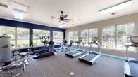 This fitness center is a comprehensively outfitted facility designed to support a full range of workout routines. The space is organized across a single floor and divided into distinct functional zones, each thoughtfully appointed with professional-grade equipment.

The cardio area is bright and inviting, lined with large windows that frame views of lush outdoor greenery and a nearby pool. Treadmills, elliptical trainers, and a stationary bike are arranged to take full advantage of the natural light and outdoor scenery. A ceiling fan overhead ensures comfortable air circulation during workouts.

Adjacent to the cardio zone, the strength training area houses an impressive array of equipment, including Cybex resistance machines, cable stations, a leg press, and a dedicated free weights section with a full dumbbell rack and kettlebells of varying weights. Rubber flooring underlays the weight equipment for safety and durability, while patterned carpet covers the broader floor area.

Architectural details such as crown molding, arched windows, and a teal accent along the base of the walls lend the space a polished, upscale character. Neutral gray walls throughout maintain a clean, focused atmosphere. A wall-mounted television is positioned for easy viewing from the strength training area, and posted fitness center rules reflect an organized, community-oriented facility.