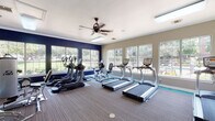 This fitness center is a comprehensively outfitted facility designed to support a full range of workout routines. The space is organized across a single floor and divided into distinct functional zones, each thoughtfully appointed with professional-grade equipment.

The cardio area is bright and inviting, lined with large windows that frame views of lush outdoor greenery and a nearby pool. Treadmills, elliptical trainers, and a stationary bike are arranged to take full advantage of the natural light and outdoor scenery. A ceiling fan overhead ensures comfortable air circulation during workouts.

Adjacent to the cardio zone, the strength training area houses an impressive array of equipment, including Cybex resistance machines, cable stations, a leg press, and a dedicated free weights section with a full dumbbell rack and kettlebells of varying weights. Rubber flooring underlays the weight equipment for safety and durability, while patterned carpet covers the broader floor area.

Architectural details such as crown molding, arched windows, and a teal accent along the base of the walls lend the space a polished, upscale character. Neutral gray walls throughout maintain a clean, focused atmosphere. A wall-mounted television is positioned for easy viewing from the strength training area, and posted fitness center rules reflect an organized, community-oriented facility.