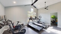 Located at 6400 Old Oak Ridge Rd in Greensboro, NC, this fitness center offers a thoughtfully designed, single-floor layout comprising four exercise rooms, a hallway, and a full bathroom. The main workout area impresses with soaring vaulted ceilings that lend a sense of openness and airiness, complemented by large multi-pane windows that flood the space with abundant natural light and offer views of surrounding greenery. Dual black ceiling fans ensure comfortable air circulation throughout the workout area. The durable speckled rubber flooring provides both safety and functionality underfoot. Equipment includes Spirit-brand treadmills, an elliptical trainer, Echelon spin bikes, and additional strength and conditioning equipment, along with exercise balls and accessories. Full-wall mirrors in a secondary exercise room visually expand the space while allowing users to monitor their form. Recessed lighting and modern ceiling fans in this mirrored room create a bright, inviting atmosphere. The private bathroom is clean and functional, featuring a single-sink vanity with white cabinetry, light wood-tone flooring, and a paper towel dispenser, making it a convenient amenity for fitness center users. The overall design is clean, modern, and purpose-built for an effective workout experience.