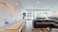 This single-floor communal lounge space is designed to foster social interaction and relaxation, blending playful aesthetics with functional amenities. The interior is anchored by a bold flamingo-print accent wall that adds a tropical, resort-inspired character throughout the main lounge area. Light wood-look flooring runs continuously across the space, contributing to a bright and cohesive feel.

The lounge is well-equipped for recreation, featuring a full-length shuffleboard table and multiple seating arrangements, including deep, comfortable sectional sofas layered over colorful geometric-patterned area rugs. A large wall-mounted flat-screen television is positioned for easy viewing from the primary seating area, while abstract artwork adorns the walls.

Floor-to-ceiling windows and glass doors line one side of the space, flooding the interior with natural light and offering unobstructed views of a resort-style outdoor pool surrounded by mature palm trees. A dark-toned dining table with contemporary chairs is positioned directly along this window wall, taking full advantage of the outdoor scenery.

The kitchenette is outfitted with white cabinetry, quartz countertops, a geometric tile backsplash, stainless steel appliances including a microwave and under-counter refrigerators, and ample storage. Recessed lighting throughout ensures the space remains well-lit at all times.