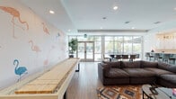 This single-floor communal lounge space is designed to foster social interaction and relaxation, blending playful aesthetics with functional amenities. The interior is anchored by a bold flamingo-print accent wall that adds a tropical, resort-inspired character throughout the main lounge area. Light wood-look flooring runs continuously across the space, contributing to a bright and cohesive feel.

The lounge is well-equipped for recreation, featuring a full-length shuffleboard table and multiple seating arrangements, including deep, comfortable sectional sofas layered over colorful geometric-patterned area rugs. A large wall-mounted flat-screen television is positioned for easy viewing from the primary seating area, while abstract artwork adorns the walls.

Floor-to-ceiling windows and glass doors line one side of the space, flooding the interior with natural light and offering unobstructed views of a resort-style outdoor pool surrounded by mature palm trees. A dark-toned dining table with contemporary chairs is positioned directly along this window wall, taking full advantage of the outdoor scenery.

The kitchenette is outfitted with white cabinetry, quartz countertops, a geometric tile backsplash, stainless steel appliances including a microwave and under-counter refrigerators, and ample storage. Recessed lighting throughout ensures the space remains well-lit at all times.
