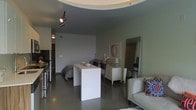 This single-floor space showcases a thoughtfully designed open-concept layout that integrates multiple living functions within one cohesive environment. A standout architectural feature is the curved, recessed ceiling element that adds a sculptural quality to the room, softening the overall aesthetic while drawing the eye upward. Track lighting mounted within this curved detail provides flexible, directed illumination throughout the space.

The polished concrete flooring runs continuously across the entire floor plan, lending an industrial yet contemporary character that anchors the design. Neutral gray walls create a calm, understated backdrop that allows furnishings and décor to take center stage.

A central white console table serves as a natural dividing element between the living and sleeping areas, which flow together without partition walls. The sleeping area features a dresser and framed artwork, while a full-length mirror enhances the sense of openness. A light-toned sofa with colorful accent pillows defines the living zone.

The kitchen, visible along one wall, is equipped with stainless steel appliances, offering functional utility integrated seamlessly into the open plan. Overall, the space conveys a modern, urban ambiance with clean lines and minimalist sensibility.