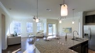 This two-story space features an open-concept layout that seamlessly connects the kitchen and living areas into one cohesive, light-filled environment. The kitchen anchors the space with a sweeping granite countertop island, finished in rich, multi-toned stone. Pendant lights hang above the island, providing both task lighting and a stylish focal point. A single-basin sink with a modern faucet completes the functional kitchen setup.

The living area extends naturally from the kitchen, showcasing dark hardwood flooring that contrasts beautifully with the light-toned walls and white ceilings. A ceiling fan with an integrated light fixture is centrally positioned over the living space, enhancing airflow and comfort. Multiple large windows along the exterior walls allow generous amounts of natural light to fill the room, contributing to its airy, open ambiance.

A sliding glass door provides direct access to an outdoor balcony, further enhancing the connection between interior and exterior. Track lighting and additional pendant fixtures ensure the space is well-lit throughout. The overall design aesthetic is modern and clean, with thoughtful lighting and quality finishes creating a polished, inviting atmosphere across both levels.