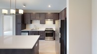 This single-floor residence offers a thoughtfully designed layout with modern finishes throughout. The kitchen serves as a central highlight, showcasing rich dark-stained cabinetry that extends to the ceiling, crisp white quartz countertops, and a full suite of stainless steel appliances including a range, dishwasher, and refrigerator. A white subway tile backsplash adds a clean, contemporary touch, while a large kitchen island provides additional prep space and casual dining. Light hardwood-style flooring runs throughout the main living areas, contributing to a warm and cohesive aesthetic. The bedroom is carpeted for comfort and features a generously sized window that frames scenic outdoor views, including glimpses of rolling hills and surrounding greenery, allowing abundant natural light to fill the room. The bathroom is well-equipped with a dark-stained vanity topped with a white countertop, a framed mirror, and a combination tub and shower outfitted with accessibility grab bars. An adjacent walk-in closet with built-in shelving and hanging rods offers excellent storage. The neutral wall tones and consistent use of dark wood cabinetry paired with white surfaces create a cohesive, modern atmosphere throughout the entire space.