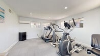 Located at 400 N 96th Ave in Tolleson, AZ, this fitness center offers a well-appointed exercise space designed to support a variety of cardio workouts. The room is finished with neutral-toned carpet tiles, crisp white walls, and recessed lighting that keeps the space bright and energetic throughout the day. Multiple windows allow natural light to fill the room while providing views of the surrounding community exterior.

The exercise room is equipped with a selection of cardio machines, including treadmills from Spirit and Life Fitness, an elliptical cross-trainer, and a stationary bike, offering users a range of workout options. A wall-mounted mini-split air conditioning unit ensures climate comfort, particularly important in the Arizona heat. A small compact refrigerator is also present for added convenience.

The entry to the fitness center features light-toned tile flooring, a large window that floods the corridor with natural light, and a decorative palm plant that adds a welcoming touch. French doors with glass panels lead directly into the exercise room, creating a clean visual connection between the entry hall and the workout space. The overall design is clean, functional, and modern, prioritizing an accessible and comfortable environment for residents seeking a convenient on-site fitness facility.