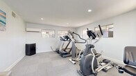 Located at 400 N 96th Ave in Tolleson, AZ, this fitness center offers a well-appointed exercise space designed to support a variety of cardio workouts. The room is finished with neutral-toned carpet tiles, crisp white walls, and recessed lighting that keeps the space bright and energetic throughout the day. Multiple windows allow natural light to fill the room while providing views of the surrounding community exterior.

The exercise room is equipped with a selection of cardio machines, including treadmills from Spirit and Life Fitness, an elliptical cross-trainer, and a stationary bike, offering users a range of workout options. A wall-mounted mini-split air conditioning unit ensures climate comfort, particularly important in the Arizona heat. A small compact refrigerator is also present for added convenience.

The entry to the fitness center features light-toned tile flooring, a large window that floods the corridor with natural light, and a decorative palm plant that adds a welcoming touch. French doors with glass panels lead directly into the exercise room, creating a clean visual connection between the entry hall and the workout space. The overall design is clean, functional, and modern, prioritizing an accessible and comfortable environment for residents seeking a convenient on-site fitness facility.