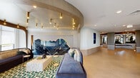 Located at 2000 Schindler Drive in South Amboy, NJ, this expansive two-floor amenity space offers a meticulously designed environment defined by upscale finishes and thoughtful layout. The main floor serves as the heart of the space, featuring a grand lobby entrance with large-format stone-look tile flooring, a decorative round mirror, and a polished credenza. A dramatic sculptural centerpiece — a tiered, curved garden planter filled with lush greenery — anchors the central atrium, which soars to double-height ceilings and is crowned by a cascading globe chandelier. A sleek staircase with brass railings and glass balustrades connects the two floors. Multiple living and lounge areas are spread across both levels, each distinctly designed. One lounge features a striking blue velvet sofa set against an organic-shaped dark marble feature wall beneath curved, wood-paneled ceilings with brass pendant lighting. Another area showcases a billiards table, built-in shelving, and a teal-accented banquette. The dining and kitchen areas are appointed with marble countertops, sage cabinetry, brass globe pendants, and mirrored backsplashes. Floor-to-ceiling windows flood the space with natural light, offering outdoor views and direct access to an exterior terrace. A two-story lounge with a fireplace and plaid-accented seating areas completes this refined, resort-style environment.