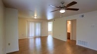 This two-story residence offers a well-organized layout with practical finishes throughout. The main living area is a highlight of the space, featuring warm hardwood flooring that reflects natural light and contributes to an inviting atmosphere. A ceiling fan with an integrated light fixture is centrally mounted on the textured ceiling, providing both airflow and supplemental lighting. Vertical blinds frame a sliding glass door, allowing natural light to filter into the room while offering privacy and potential outdoor access.

The living area connects to additional spaces visible through interior doorways. One passage leads to what appears to be a secondary room or hallway, illuminated by a flush-mount ceiling light, suggesting a functional flow between spaces. To the right, a partial view reveals a bathroom, identifiable by its warm-toned cabinetry and ambient lighting, indicating convenient proximity to the main living area.

The neutral wall tones and light-colored trim throughout create a clean, cohesive aesthetic. With two floors of living space, this home offers room for varied uses, balancing open communal areas with more private spaces distributed across its multi-level layout.