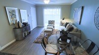 This single-floor space offers a clean and functional layout with a distinctly bright and neutral aesthetic throughout. The kitchen is a standout feature, designed with an open-concept approach and separated from the adjacent living area by a peninsula-style breakfast bar topped with a light speckled countertop — ideal for casual dining or meal prep. The kitchen is outfitted with ample white upper and lower cabinetry, providing generous storage, and is equipped with a white electric range, a matching refrigerator, and an under-cabinet range hood. Overhead, a flush fluorescent light fixture ensures the workspace is well-illuminated. The uniformly white cabinetry and appliances, paired with the light-toned walls and countertops, create a cohesive and airy atmosphere. The neutral color scheme — predominantly whites and soft grays — lends a timeless and versatile quality to the interior, making it easy to personalize. The single-floor plan ensures convenient, step-free living throughout. Overall, the space presents a tidy, move-in ready environment with a straightforward, practical design focused on comfort and functionality.