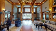 Located at 15155 Richmond Ave in Houston, TX, this single-story clubhouse lobby is a thoughtfully designed communal space that combines warmth and sophistication. The grand entry immediately impresses with soaring double-height ceilings framed by rich warm wood millwork and structural columns, creating a sense of openness and architectural grandeur.

The main lounge area features a sleek, modern fireplace flanked by plush blue velvet armchairs and a stone accent wall that rises dramatically toward the ceiling. Large floor-to-ceiling windows and glass panels flood the space with natural light and offer views of the outdoor pool area beyond, seamlessly connecting the interior to the exterior amenities.

Polished concrete and natural stone tile flooring runs throughout, adding a grounded, earthy contrast to the refined woodwork above. A generous sectional sofa anchors a secondary seating area beneath a built-in wood shelving display unit, providing a comfortable and inviting space for residents to gather.

Glazed interior doors with wood transoms lead to additional amenities, including a fitness center and media room, clearly marked for resident use. Wall sconces provide warm ambient lighting throughout, complementing the recessed ceiling fixtures. The overall ambiance is upscale yet welcoming, blending craftsman-inspired architectural details with contemporary furnishings for a refined communal experience.