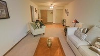 This single-floor residence offers a comfortable and functional layout designed for everyday living. The living room serves as the heart of the space, featuring warm neutral wall tones and a combination of carpet and hardwood-style flooring that define distinct areas within the room. A ceiling fan with integrated lighting provides both air circulation and ambient illumination overhead. The living area is furnished with a plush sofa and an accent armchair dressed with decorative green pillows, centered around a wooden coffee table that anchors the seating arrangement. A baseboard heating unit runs along the wall, ensuring year-round comfort. Greenery in the form of a tall potted plant adds a natural, refreshing touch to the room's ambiance. Toward the back of the living space, a sliding closet with wooden panel doors offers generous storage, while a solid entry door with a decorative mat provides a welcoming transition point. Wall-mounted photo frames add a personalized, homey character to the space. A glimpse into an adjacent area reveals additional furnishings, suggesting a connected dining or flex space. Overall, the residence presents a warm, lived-in atmosphere with practical features throughout.