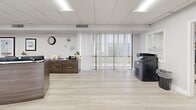 Located at 2903-2915 NW 60th Terr in Sunrise, Florida, this well-appointed single-floor commercial space offers a clean, modern aesthetic designed for professional use. The layout flows seamlessly from a bright, open reception and waiting area near the entrance — furnished with sleek black leather seating, a marble-top accent table, and wall-mounted displays — to a curved, dark wood reception desk that serves as the functional centerpiece of the space. Light wood-look flooring runs throughout, complementing the crisp white walls and drop-panel ceiling with recessed and panel lighting that keeps the entire space evenly illuminated. Large windows and sliding glass doors at both ends of the suite allow generous natural light to pour in and provide views of the surrounding outdoor deck and landscaping. The kitchen area features a white peninsula counter with bar-height stools, ideal for breaks or informal meetings. Two private offices offer dedicated workspaces, while a closet provides additional storage. The overall ambiance is polished and professional, with a neutral color palette and thoughtful layout that balances an open, collaborative feel with practical privacy — making this a versatile and functional commercial environment.