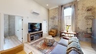 Located at 745 E Main St in Chattanooga, TN, this 2-bedroom, 1-bathroom unit artfully combines historic industrial character with contemporary design. The open-concept living and kitchen area is the centerpiece of the space, featuring soaring ceilings, exposed brick and distressed plaster walls, and tall windows that flood the room with natural light. Light wood flooring runs throughout, complementing the warm tones of the brick. The kitchen is equipped with stainless steel appliances, including a full-size refrigerator and range, and features gray cabinetry with a clean white countertop. A wall-mounted mini-split unit provides climate control. The primary bedroom retains the building's historic character with striking exposed brick and weathered plaster walls, a small window offering a green outdoor view, and light hardwood flooring. The second bedroom contrasts with crisp white walls, a built-in closet with paneled doors, and a calm, neutral atmosphere. The shared bathroom is neatly appointed with a white vanity, vessel sink, industrial-style light fixtures, a large mirror, and a tub-shower combination. Throughout the unit, the interplay of raw historic materials and clean modern updates creates a distinctive urban aesthetic.
