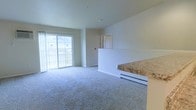 This single-floor space features an open-concept layout that seamlessly connects the living area to the kitchen. The living room is finished with wall-to-wall carpeting in a neutral gray tone, creating a comfortable and cohesive feel throughout. A large sliding glass door dominates one wall, flooding the space with natural light and providing direct access to an outdoor balcony, where views of the surrounding area are visible. A baseboard heating unit runs along the exterior wall, ensuring efficient climate control.

The kitchen is partially visible through a half-wall pass-through with a granite-style countertop, offering a functional connection between the cooking and living spaces while maintaining a degree of separation. Adjacent to the living area, a doorway leads to additional rooms within the unit. The walls are finished in a soft, neutral tone that complements the light carpeting and contributes to the overall bright and airy ambiance. The layout is practical and straightforward, making efficient use of the available space while maintaining an open, uncluttered atmosphere throughout.