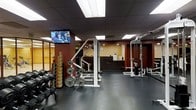 This fitness center is a fully appointed single-floor facility designed to meet a wide range of strength and conditioning needs. The space features a dark rubber floor that provides both durability and safety, ideal for heavy equipment use. A variety of free weights are prominently available, including an organized dumbbell rack with a full selection of weights, as well as plate-loaded barbells and weight stacks. Strength training machines, including a Smith machine and a cable/pulley rack system, are centrally positioned, offering users versatile workout options. The drop ceiling is fitted with recessed fluorescent panel lighting that evenly illuminates the entire floor, creating a bright and functional workout environment. A wall-mounted flat-screen television is positioned for easy viewing during workouts. The warm, earthy tones of the walls — featuring deep red and tan accents — contribute to an energizing atmosphere. Toward the rear of the space, a staffed reception or front desk area is visible, suggesting professional management and facility oversight. The overall layout is open and efficiently organized, allowing for comfortable movement between equipment stations.
