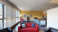 This single-floor urban space combines modern design with a lively, colorful aesthetic. The open-concept layout seamlessly connects the living area and kitchen, creating a sociable and functional environment. Bold yellow accent walls energize the space and provide a warm backdrop throughout.

The living area is generously sized, featuring large floor-to-ceiling windows that flood the room with natural light and offer views of the surrounding cityscape. Exposed industrial-style ductwork runs along the upper walls, lending a contemporary loft character to the space. Dark polished concrete floors ground the interior with a sleek, urban feel.

The kitchen is efficiently designed with light maple cabinetry, dark granite countertops, stainless steel appliances, and a classic subway tile backsplash. A bar-style peninsula with quilted metallic paneling and silver bar stools serves as both a prep area and casual dining spot, illuminated by stylish pendant lights overhead.

The bathroom continues the cohesive design language, featuring yellow walls, a vessel sink set atop a maple vanity with dark granite countertops, a large mirror, and stone-look tile flooring.

Overall, the space presents a thoughtfully designed, urban-inspired interior that balances bold color choices with modern fixtures and an abundance of natural light.