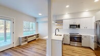 This single-floor space showcases a clean, contemporary design defined by light wood-look flooring and bright white walls that create a cohesive, airy atmosphere throughout. The open-concept kitchen is a central highlight, featuring white flat-panel cabinetry, a white quartz peninsula with an undermount sink and matte black faucet, and a full suite of stainless steel appliances including a refrigerator, range, and built-in microwave. Warm-toned live-edge floating shelves and pendant lighting add character above the peninsula, while a white subway tile backsplash completes the polished look.

The living area flows seamlessly from the kitchen and is bathed in natural light from a set of French doors that open to an outdoor green space, as well as a large window fitted with horizontal blinds. A built-in low shelving unit sits beneath the window, offering practical storage. Recessed lighting supplements natural light throughout.

The bedroom is generously proportioned with a window overlooking the surroundings and access to a well-organized walk-in closet with built-in shelving. The bathroom maintains the same modern aesthetic with white cabinetry, a wide vanity mirror, and matte black hardware accents, all set against the consistent wood-look flooring.