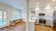 This single-floor space showcases a clean, contemporary design defined by light wood-look flooring and bright white walls that create a cohesive, airy atmosphere throughout. The open-concept kitchen is a central highlight, featuring white flat-panel cabinetry, a white quartz peninsula with an undermount sink and matte black faucet, and a full suite of stainless steel appliances including a refrigerator, range, and built-in microwave. Warm-toned live-edge floating shelves and pendant lighting add character above the peninsula, while a white subway tile backsplash completes the polished look.

The living area flows seamlessly from the kitchen and is bathed in natural light from a set of French doors that open to an outdoor green space, as well as a large window fitted with horizontal blinds. A built-in low shelving unit sits beneath the window, offering practical storage. Recessed lighting supplements natural light throughout.

The bedroom is generously proportioned with a window overlooking the surroundings and access to a well-organized walk-in closet with built-in shelving. The bathroom maintains the same modern aesthetic with white cabinetry, a wide vanity mirror, and matte black hardware accents, all set against the consistent wood-look flooring.
