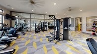 This single-floor amenity space is thoughtfully designed to serve both fitness enthusiasts and families, combining a well-appointed fitness center with a dedicated children's play area. The two zones are connected by a corridor and separated by glass partition walls, allowing visual continuity while maintaining distinct functions.

The fitness center is the centerpiece of the space, offering a generous open layout filled with a comprehensive selection of equipment. Cybex cable machines, treadmills, ellipticals, stationary bikes, rowing machines, and a full dumbbell rack are arranged to allow comfortable circulation. Large windows line multiple walls, inviting abundant natural light and providing outdoor views of the surrounding property. Ceiling fans and recessed lighting contribute to a well-ventilated, bright atmosphere. Full-length mirrors along one wall visually expand the room and aid in form monitoring.

Adjacent to the gym, the children's play room features playful robot-themed wall decals, a wall-mounted flat-screen television, a low media console with storage cubbies, bean bag seating, and interactive tablet stations. Large LEGO-inspired decorative elements add a whimsical, imaginative touch.

Throughout both areas, the signature geometric yellow, gray, and white patterned carpet ties the entire space together with a cohesive, energetic aesthetic.