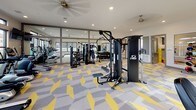 This single-floor amenity space is thoughtfully designed to serve both fitness enthusiasts and families, combining a well-appointed fitness center with a dedicated children's play area. The two zones are connected by a corridor and separated by glass partition walls, allowing visual continuity while maintaining distinct functions.

The fitness center is the centerpiece of the space, offering a generous open layout filled with a comprehensive selection of equipment. Cybex cable machines, treadmills, ellipticals, stationary bikes, rowing machines, and a full dumbbell rack are arranged to allow comfortable circulation. Large windows line multiple walls, inviting abundant natural light and providing outdoor views of the surrounding property. Ceiling fans and recessed lighting contribute to a well-ventilated, bright atmosphere. Full-length mirrors along one wall visually expand the room and aid in form monitoring.

Adjacent to the gym, the children's play room features playful robot-themed wall decals, a wall-mounted flat-screen television, a low media console with storage cubbies, bean bag seating, and interactive tablet stations. Large LEGO-inspired decorative elements add a whimsical, imaginative touch.

Throughout both areas, the signature geometric yellow, gray, and white patterned carpet ties the entire space together with a cohesive, energetic aesthetic.