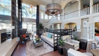 This impressive communal space makes an immediate statement with its soaring double-height ceilings and an abundance of natural light streaming through floor-to-ceiling windows that frame views of lush palm trees beyond. A large, industrial-style drum chandelier serves as a dramatic focal point, anchoring the main seating area below with warmth and sophistication.

The main lounge area is thoughtfully arranged with plush, neutral-toned sofas and accent chairs accented by colorful throw pillows, all set atop a generous area rug over rich hardwood flooring. A sleek glass-top coffee table and dark wood partitions help define the seating zones while maintaining an open, airy feel throughout.

An elegant curved staircase with white balusters leads to an open upper-level mezzanine, lined with a dark wood railing that overlooks the lounge below. Arched architectural details and built-in shelving add classic charm to the upper level. Lush indoor plants are thoughtfully placed throughout, adding vibrancy and a connection to the natural surroundings. The overall ambiance is refined yet inviting, combining upscale design elements with a comfortable, resort-inspired atmosphere.