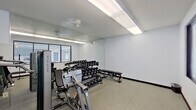 This single-floor fitness room offers a functional and well-organized workout environment designed to support a range of strength training activities. The space features durable rubber tile flooring throughout, providing cushioning and grip suitable for free weight and machine-based exercises. Large windows along one wall allow natural light to enter and offer views of what appears to be an outdoor pool area, lending an open, airy quality to the room.

A floor-to-ceiling mirror spans a significant portion of one wall, visually expanding the space and allowing users to monitor their form during workouts. The room is equipped with Matrix-branded strength machines, including a cable/functional trainer and a selectorized weight machine, positioned to make efficient use of the available floor area. A multi-tier dumbbell rack holds a comprehensive range of free weights, accompanied by a set of kettlebells and adjustable workout benches. Overhead fluorescent lighting ensures consistent, bright illumination throughout the space. The room is accessed via a standard commercial-grade door with a window panel, and safety features include a clearly marked exit sign and a fire alarm. The overall aesthetic is clean and utilitarian, prioritizing practicality and functionality.