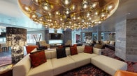 This elegantly designed single-floor amenity space makes a bold statement with its interior design and carefully curated furnishings. The centerpiece of the main lounge is a dramatic ceiling installation of clustered gold and smoked-glass spherical pendant lights, casting a warm, ambient glow over a generously sized sectional sofa dressed in rich crimson and charcoal throw pillows. Bold, abstract patterned rugs in deep reds and earth tones anchor the various seating arrangements throughout the space.

Multiple distinct zones offer a variety of uses. A curved leather sofa grouping provides an intimate conversation area, while a secluded nook features a round pedestal table surrounded by high-backed upholstered chairs in soft grey with gold-toned bases, spotlit to highlight a large floral art piece on the wall. Stone-effect wall cladding runs consistently throughout, lending a cohesive, contemporary texture to the interior.

The kitchenette area is appointed with sleek dark wood cabinetry, quartz countertops, a stainless steel sink with a brass faucet, a built-in oven, and dual wall-mounted flat-screen televisions. Wide-plank hardwood flooring and a wood-paneled ceiling complete the refined aesthetic, while large windows allow natural light to filter in and provide views to the exterior surroundings.