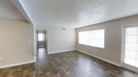 This single-floor space presents a straightforward and functional layout designed for comfortable everyday living. The main living area is finished with durable wood-style plank flooring in a warm, mid-toned hue that extends throughout the space, providing a cohesive and easy-to-maintain surface underfoot. Neutral, light gray walls keep the interior feeling open and versatile, complementing a variety of furnishing styles.

The ceiling features a classic textured popcorn finish, adding a layer of visual depth overhead. A generously sized window along one wall allows natural light to fill the room, enhancing the sense of openness and warmth. A visible HVAC vent indicates a central air system for year-round climate control.

A hallway visible from the main living area leads toward additional rooms, hinting at a practical and efficient floor plan. The corridor is brightened by natural light at its far end, suggesting exterior access or a windowed room beyond. White baseboards frame the flooring neatly throughout, adding a clean, finished look to the overall interior. The space is move-in ready, presenting a neutral canvas well-suited for personalization.